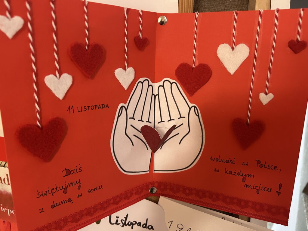 w środku czerwonej kartki znajdują się serduszka oraz w dłoniach serce i napis: 11 listopada dziś świętujemy z dumą w sercu wolność w Polsce w każdym miejscu