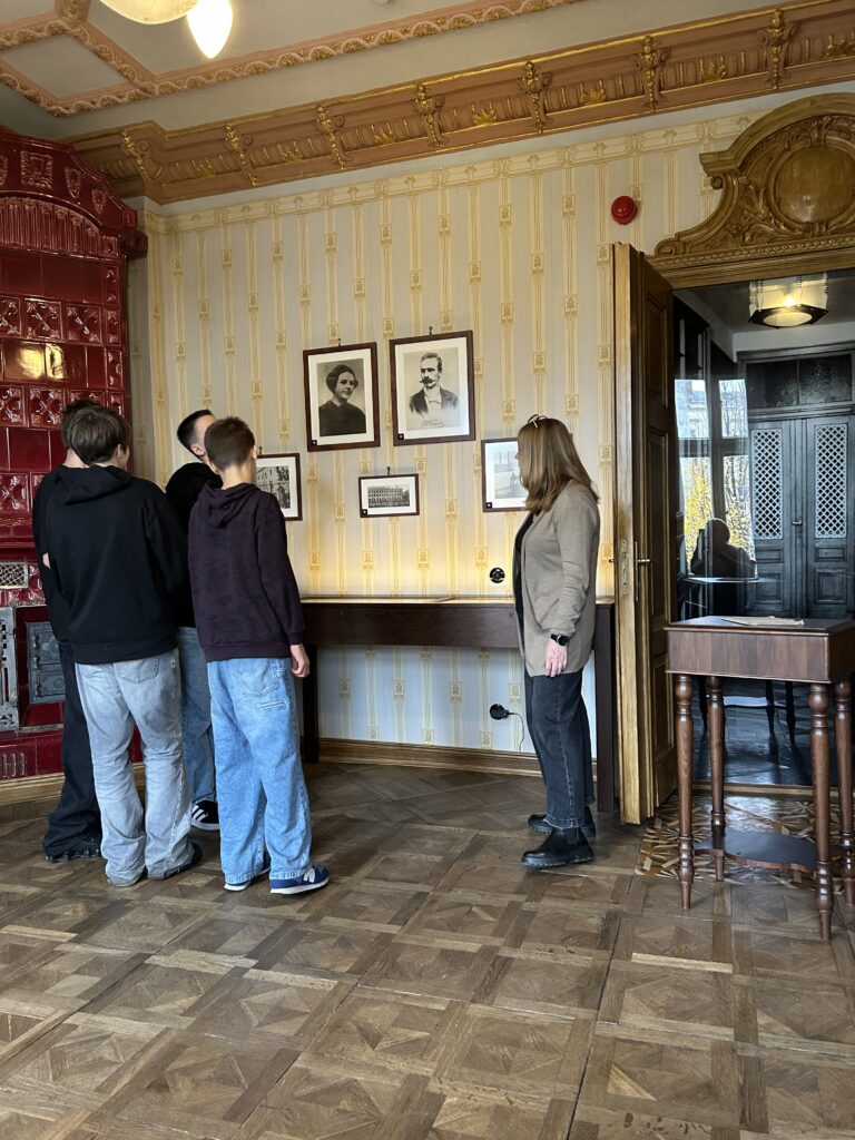 chłopcy oglądają zdjęcia wiszące na ścianie przedstawiające Józefa Piłsudskiego oraz jego małżonkę. Pani opowiada im, co przedstawiają zdjęcia. Chłopcy stoją tyłem do aparatu