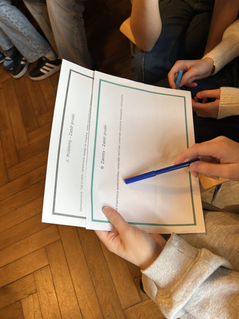 na zdjęciu dwie karty pracy z napisem: zasoby - zabór pruski, problemy - zabór pruski. Widać także dłonie i mazak, którym są wskazywane słowa