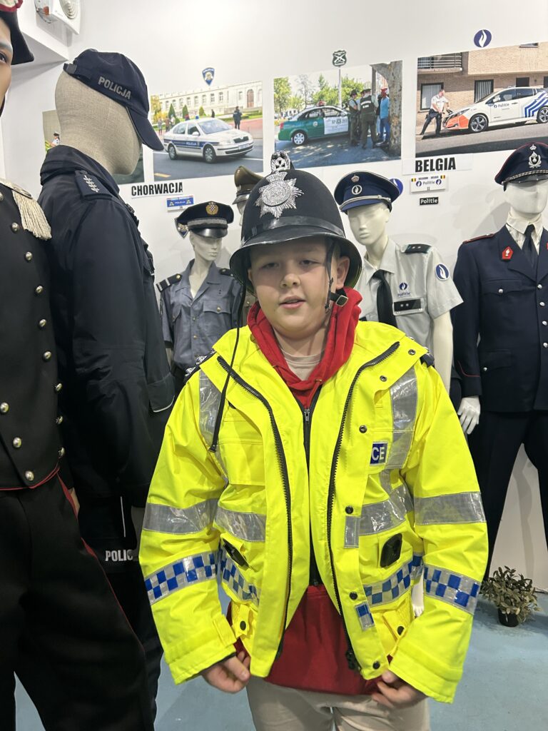 na pierwszym planie chłopiec ubrany w odblaskową, jasnozieloną kurtkę oraz hełm policjanta z Wysp Brytyjskich