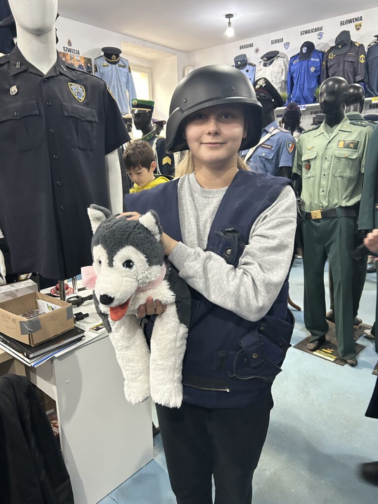 na pierwszym planie dziewczynka ubrana w czarny hełm bojowy oraz granatową kamizelką. Pod pachą trzyma dużego. pluszowego psa