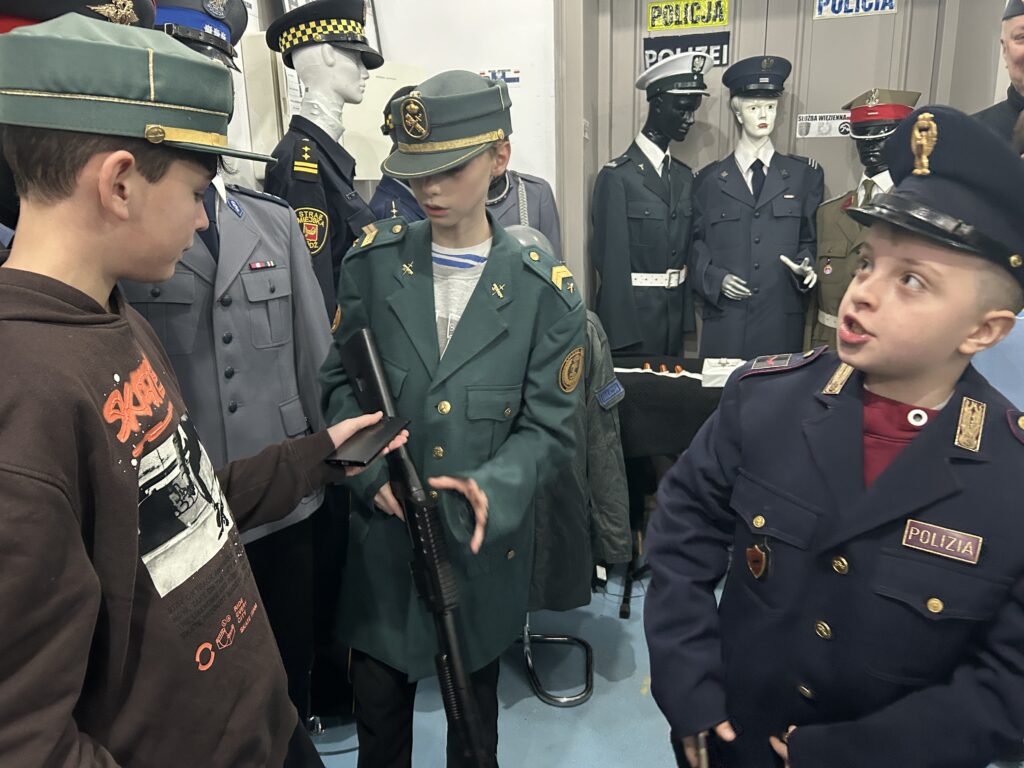 na pierwszym planie trzej chłopcy. jeden ubrany w czapkę policyjną, drugi i trzeci także w mundury z różnych państw. Jeden w ręku trzyma zabawkowy karabun skierowany do dołu. Na drugim planie manekiny w mundurach polskich milicjantów i współczesnej policji