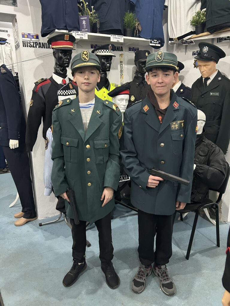 na pierwszym planie dwaj chłopcy w mundurach i czapkach policyjnych w kolorze ciemnozielonym. Jeden z chłopców trzyma w ręku zabawkowy pistolet