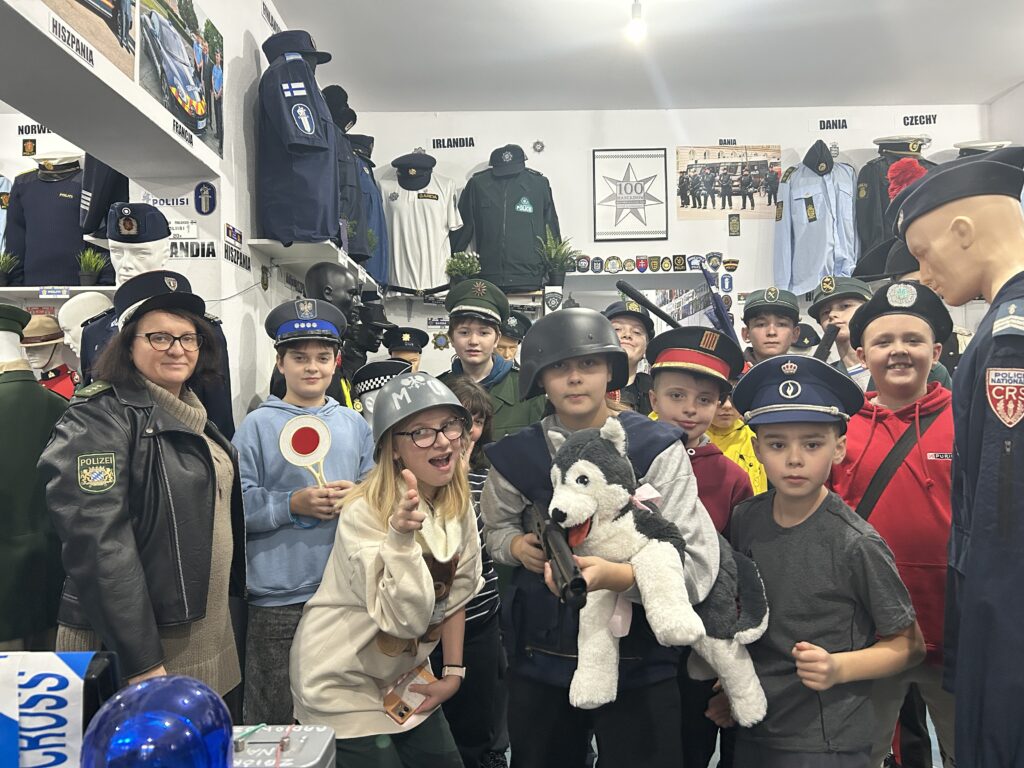 zdjęcie grupowe - dzieci w muzeum mundurów na tle manekinów. Dzieci ubrane w różne mundury i czapki. Po lewej stronie zdjęcia stoi także pani w kurtce i czapce policyjnej