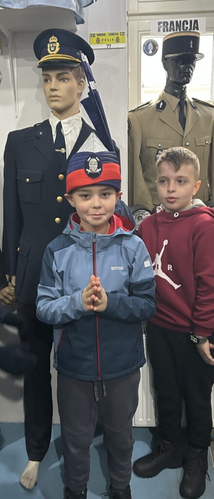 dwaj chłopcy na pierwszym planie, za nimi  dwa manekiny w mundurach. Jeden z chłopców ma paradną czapkę policyjną z kraju azjatyckiego