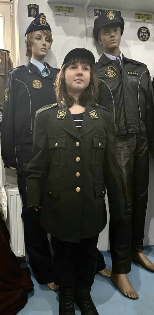 na tle dwóch manekinów stoi na baczność dziewczynka w ciemnozielonym mundurze i czapce policyjnej
