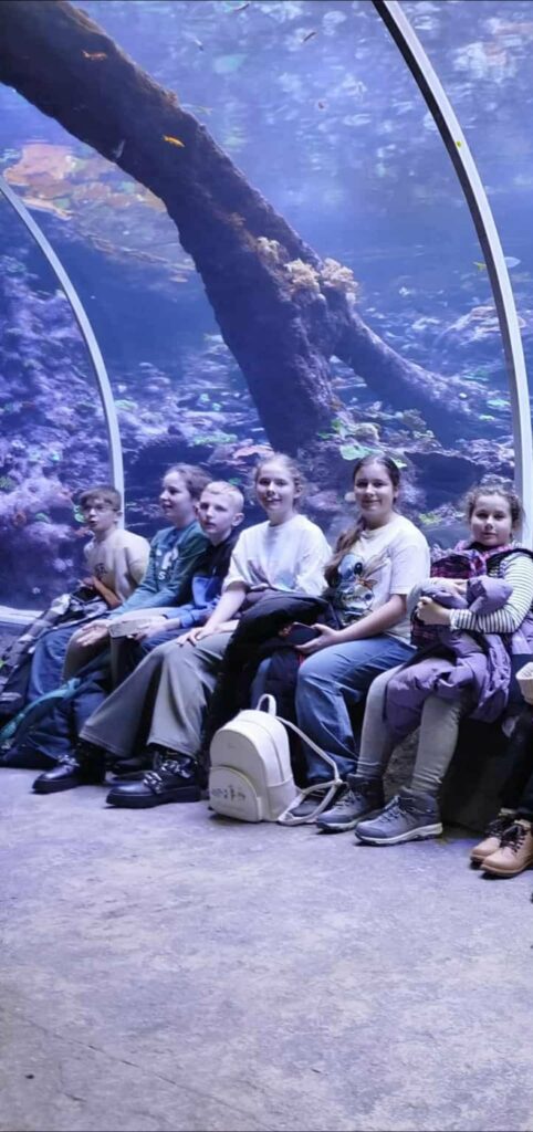 zdjęcie grupowe uczniów siedzących w orientarium, nad nimi akwarium 