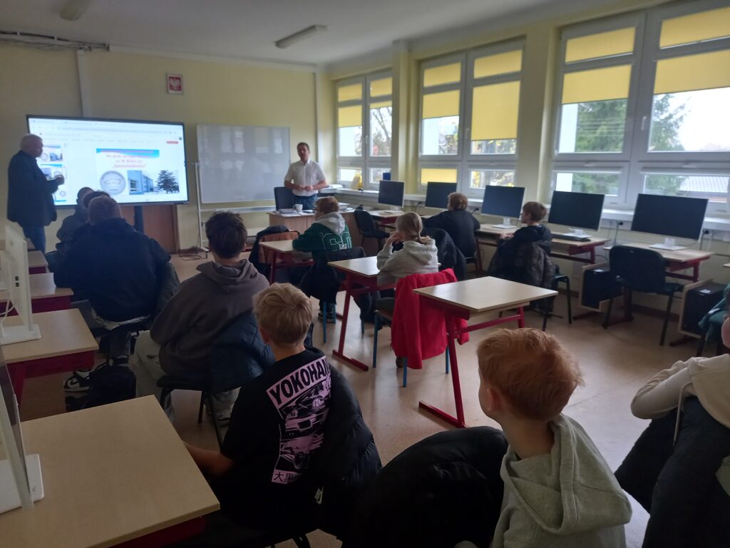 dzieci siedząc w ławkach słuchają wykładu, na końcu na ekranie interaktywnym są wyświetlane slajdy