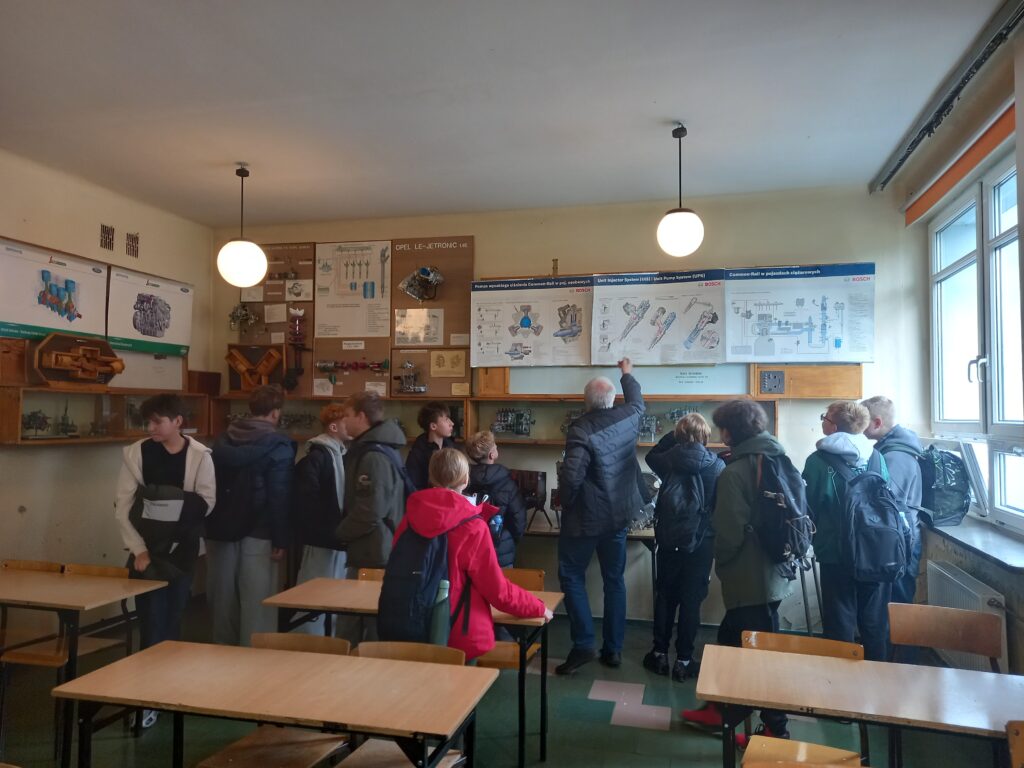 Dzieci stoją w klasie, mężczyzna tłumaczy im działanie pewnych mechanizmów, pokazując elementy na plakatach powieszonych u góry