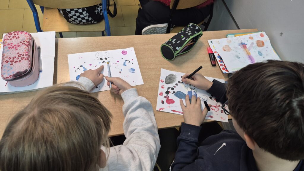 Uczniowie pochyleni nad ławką robią kleksy na białych kartkach