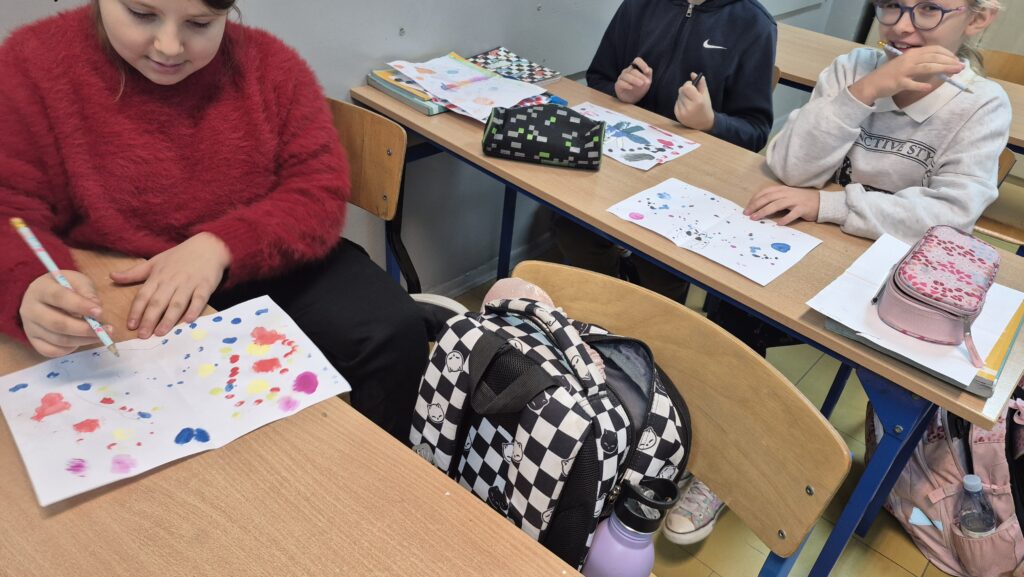 Uczniowie siedzą w ławkach i robią kleksy na białych kartkach