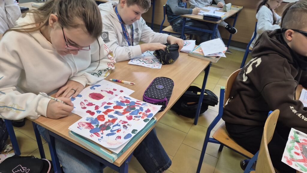 Uczniowie siedzą w ławkach i robią kleksy na białych kartkach