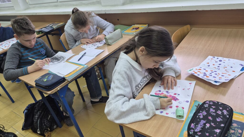 Uczennice siedzą w ławkach i robią kleksy na białych kartkach