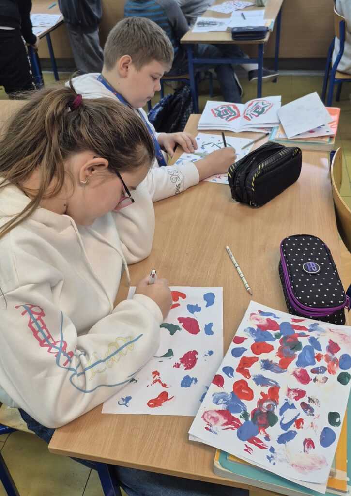 Uczniowie siedzą w ławkach i robią kleksy na białych kartkach