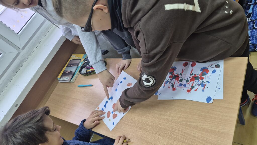 Uczniowie pochyleni nad ławką robią kleksy na białych kartkach