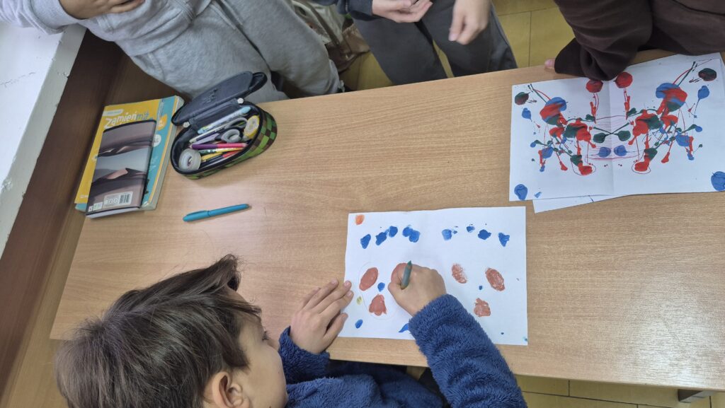 Uczniowie pochyleni nad ławką robią kleksy na białych kartkach