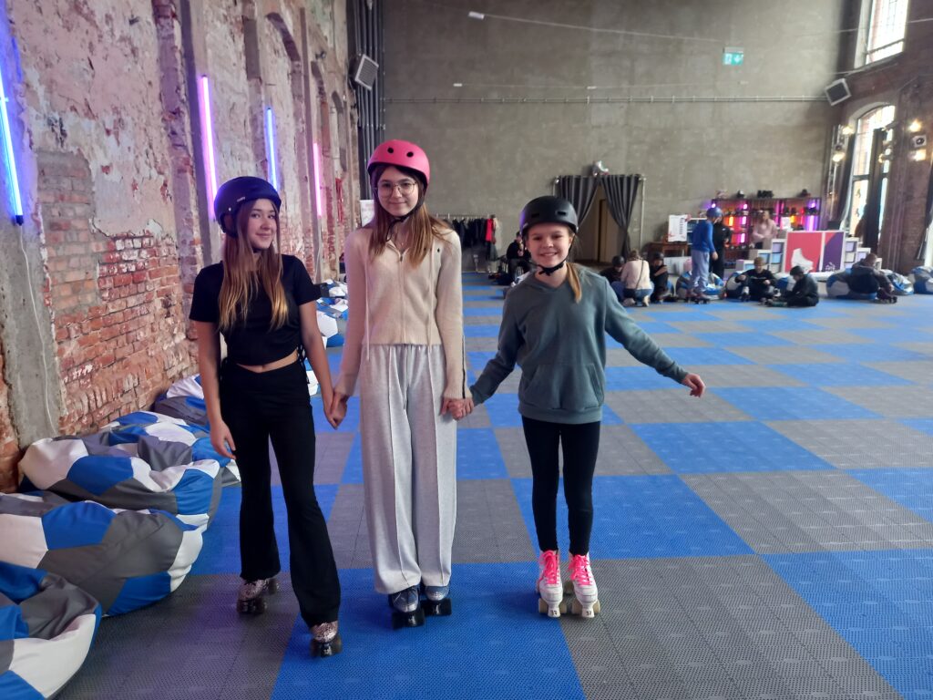 Trzy uczennice mają wrotki i kaski, stoją razem. 