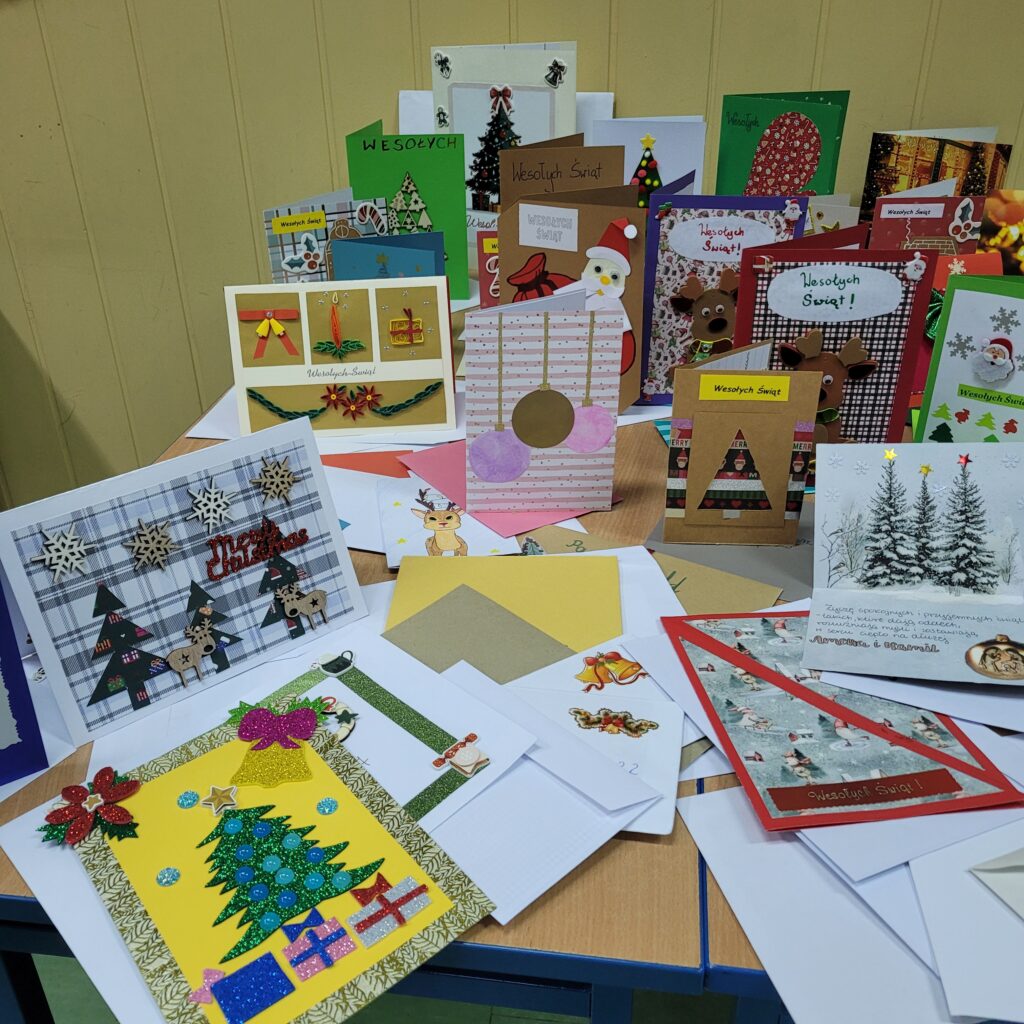 zdjęcie ławki na której stoją wykonane ręcznie kartki świąteczne przez uczniów. Są kolorowe w kolorach zieleni, czerwieni, złota, niebieskiego, w choinki, prezenty, krajobrazy itp. Część w kopertach. 
