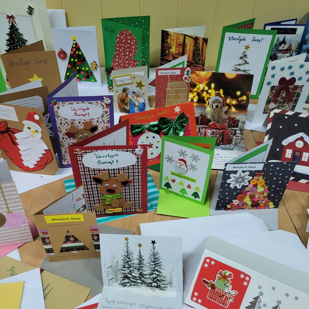 zdjęcie ławki na której stoją wykonane ręcznie kartki świąteczne przez uczniów. Są kolorowe w kolorach zieleni, czerwieni, złota, niebieskiego, w choinki, prezenty, krajobrazy itp. Część w kopertach. 