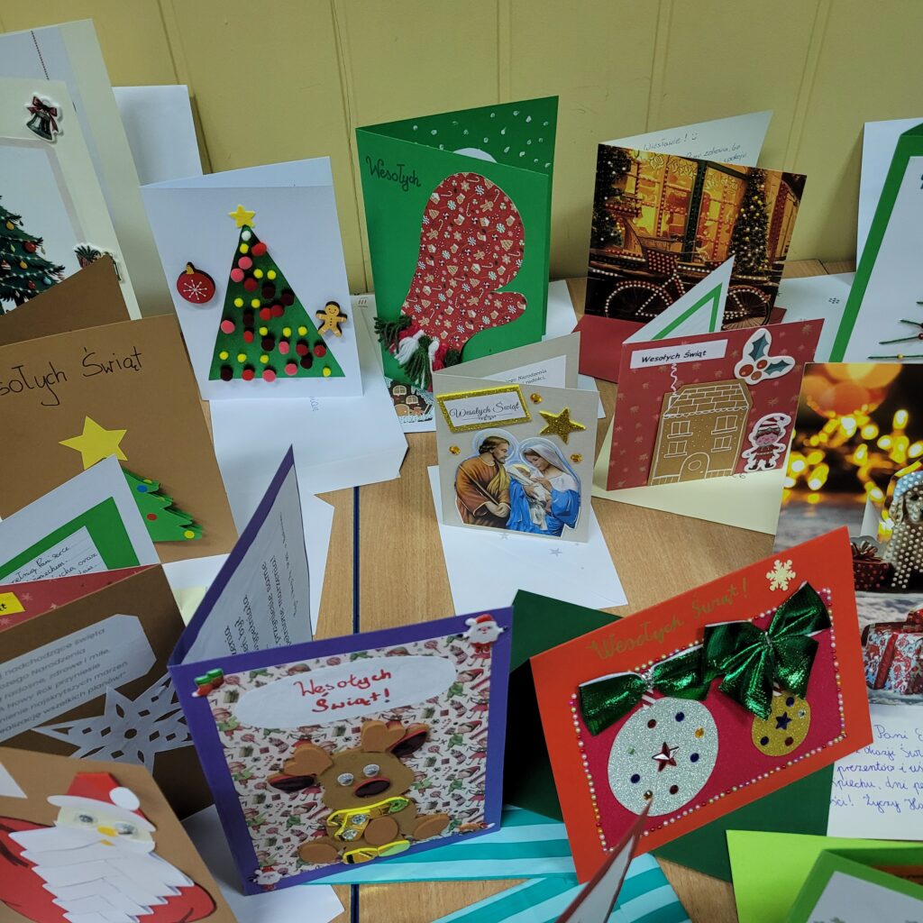 zdjęcie ławki na której stoją wykonane ręcznie kartki świąteczne przez uczniów. Są kolorowe w kolorach zieleni, czerwieni, złota, niebieskiego, w choinki, prezenty, krajobrazy itp. Część w kopertach. 
