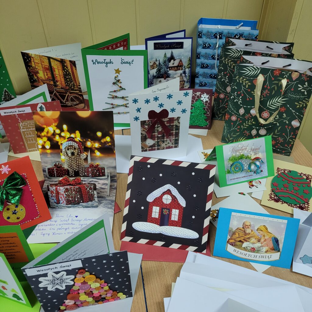 zdjęcie ławki na której stoją wykonane ręcznie kartki świąteczne przez uczniów. Są kolorowe w kolorach zieleni, czerwieni, złota, niebieskiego, w choinki, prezenty, krajobrazy itp. Część w kopertach. 