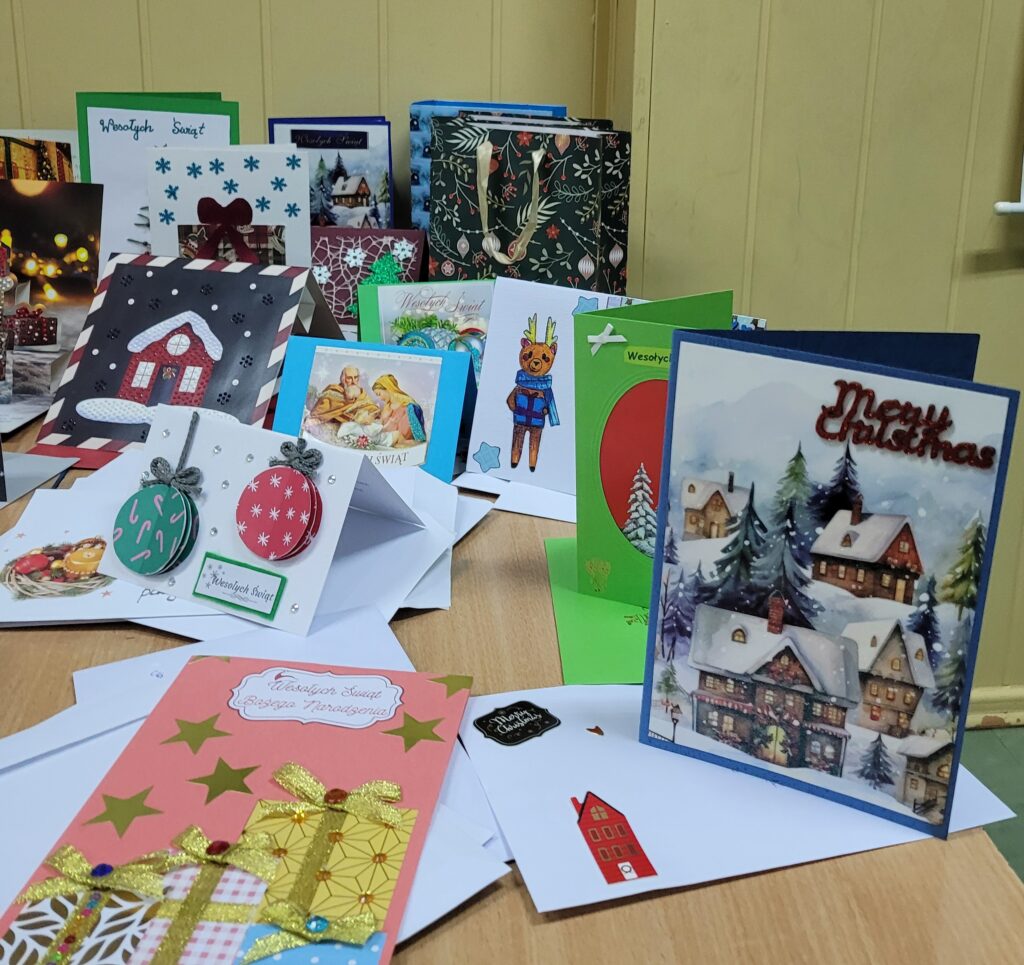 zdjęcie ławki na której stoją wykonane ręcznie kartki świąteczne przez uczniów. Są kolorowe w kolorach zieleni, czerwieni, złota, niebieskiego, w choinki, prezenty, krajobrazy itp. Część w kopertach. 