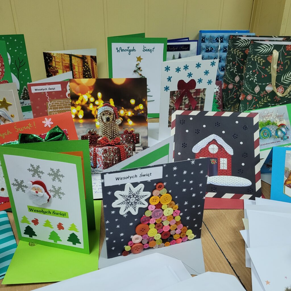 zdjęcie ławki na której stoją wykonane ręcznie kartki świąteczne przez uczniów. Są kolorowe w kolorach zieleni, czerwieni, złota, niebieskiego, w choinki, prezenty, krajobrazy itp. Część w kopertach. 