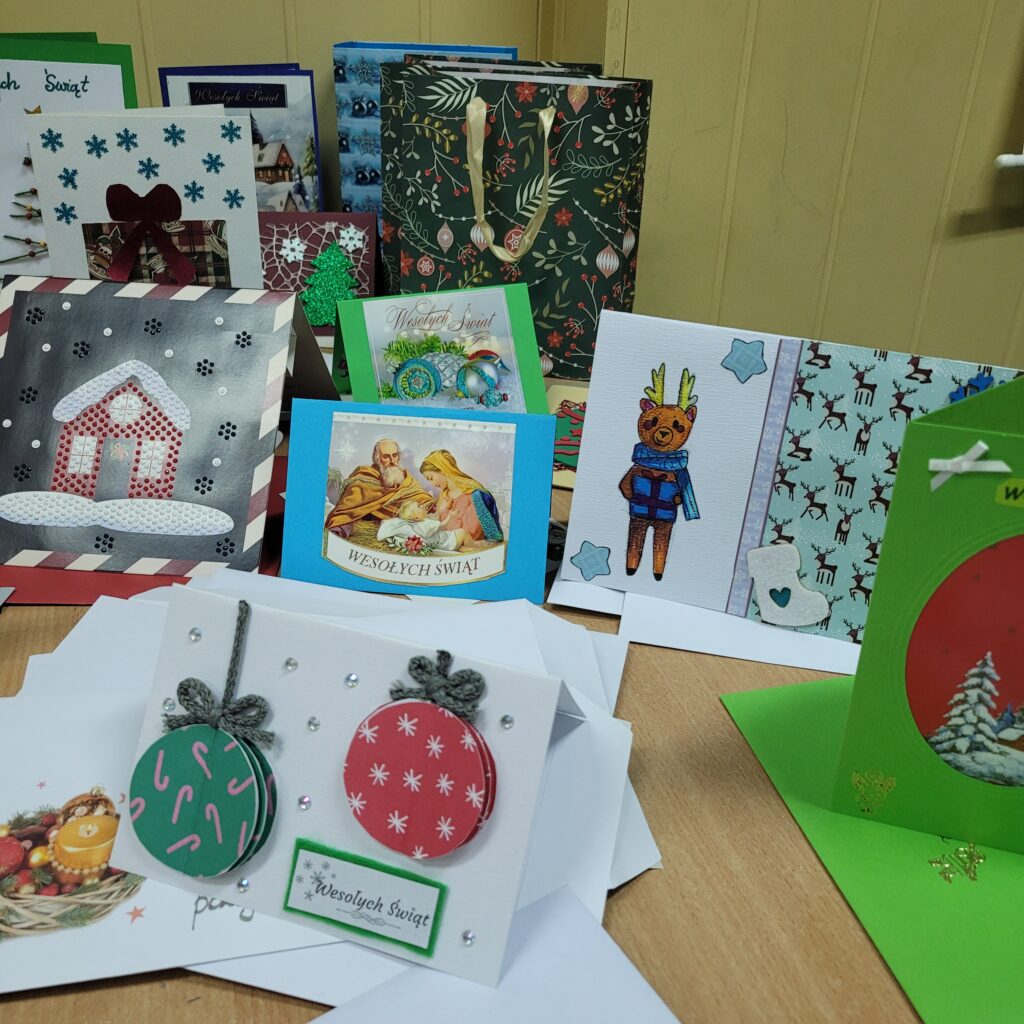 zdjęcie ławki na której stoją wykonane ręcznie kartki świąteczne przez uczniów. Są kolorowe w kolorach zieleni, czerwieni, złota, niebieskiego, w choinki, prezenty, krajobrazy itp. Część w kopertach. 