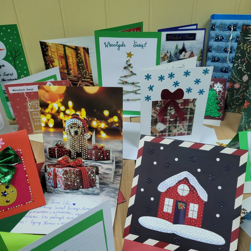 zdjęcie ławki na której stoją wykonane ręcznie kartki świąteczne przez uczniów. Są kolorowe w kolorach zieleni, czerwieni, złota, niebieskiego, w choinki, prezenty, krajobrazy itp. Część w kopertach. 