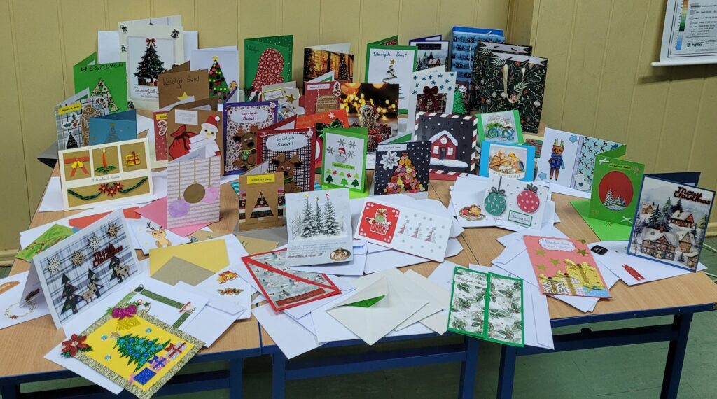 zdjęcie ławki na której stoją wykonane ręcznie kartki świąteczne przez uczniów. Są kolorowe w kolorach zieleni, czerwieni, złota, niebieskiego, w choinki, prezenty, krajobrazy itp. Część w kopertach. 
