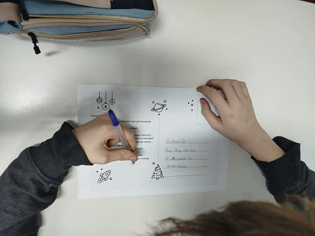 Na zdjęciu widoczne są dłonie ucznia podpisującego kartkę świąteczną do Prezesa Rady Ministrów.
