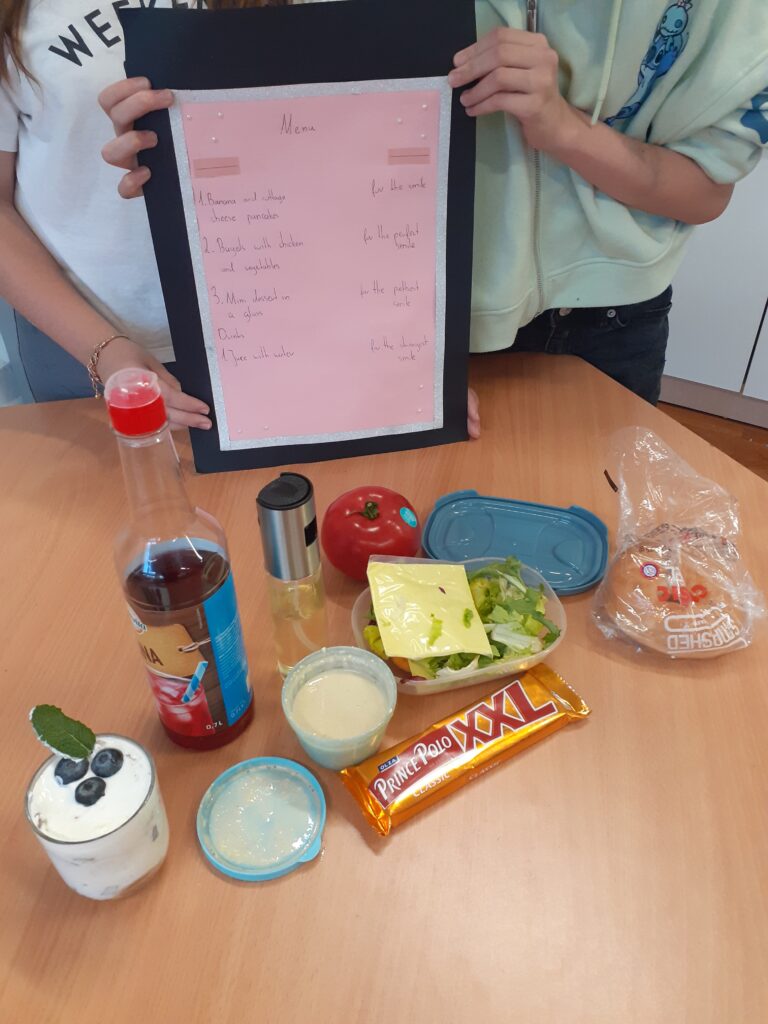 na zdjęciu widać wyłożone na stole składniki potrzebne do przygotowania potraw oraz plakat w języku angielskim, trzymany przez autorki przepisów.