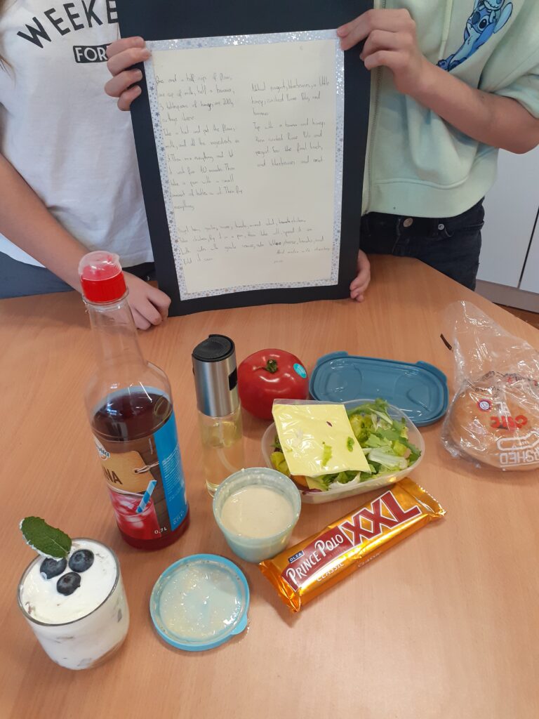 na zdjęciu widać wyłożone na stole składniki potrzebne do przygotowania potraw oraz plakat w języku angielskim, trzymany przez autorki przepisów.