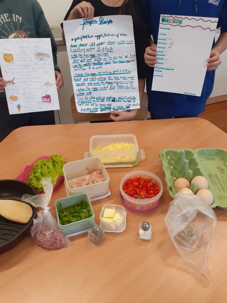 na zdjęciu widać wyłożone na stole składniki potrzebne do przygotowania omletów warzywnych oraz plakaty w języku angielskim, trzymane przez autorki przepisów.