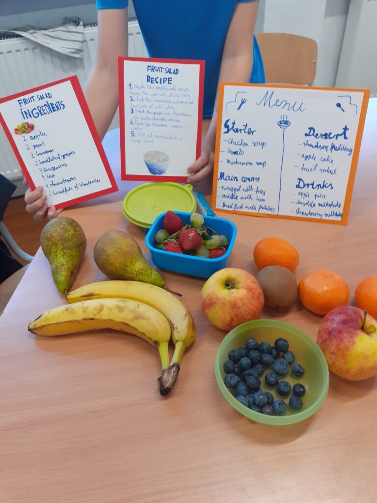 na zdjęciu widać wyłożone na stole owoce potrzebne do przygotowania sałatki owocowej oraz wyeksponowane plakaty w języku angielskim z przepisem na sałatkę oraz propozycją menu szkolnej stołówki.