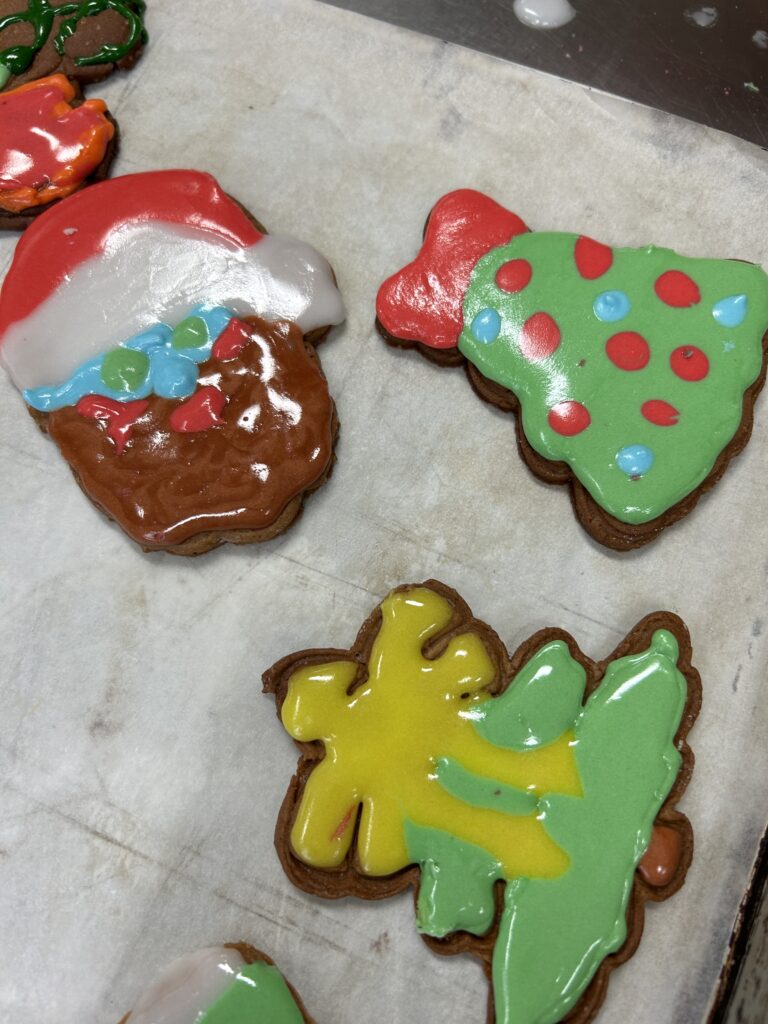 na stole leżą trzy pierniczki - jeden to głowa Mikołaja pomalowany kolorowymbiało-czerwono-niebieskim lukrem, drugi to dzwoneczek pomalowany zielonym lukrem, na nim niebiesko-czerwone kropeczki i na górze na czerwono pomalowana kokardka. Trzecie ciateczko jest w kształcie choinki pomalowane lukrem zielonym i żółtym