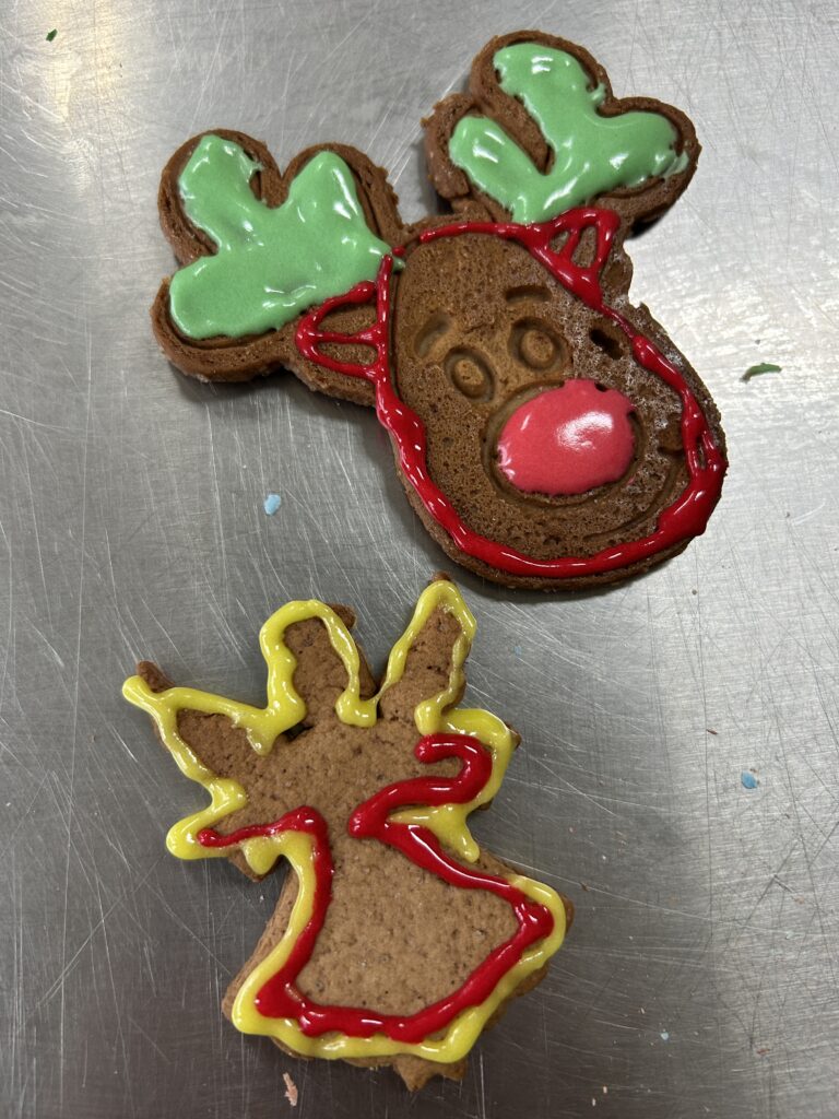 na stole leżą dwa pierniczki - jeden w kształcie głowy reniferaz czerwonym noskim i zielonym porożem. Drugie-w kształcie aniołka udekorowane, obrysowane czerwono-żółtym lukrem