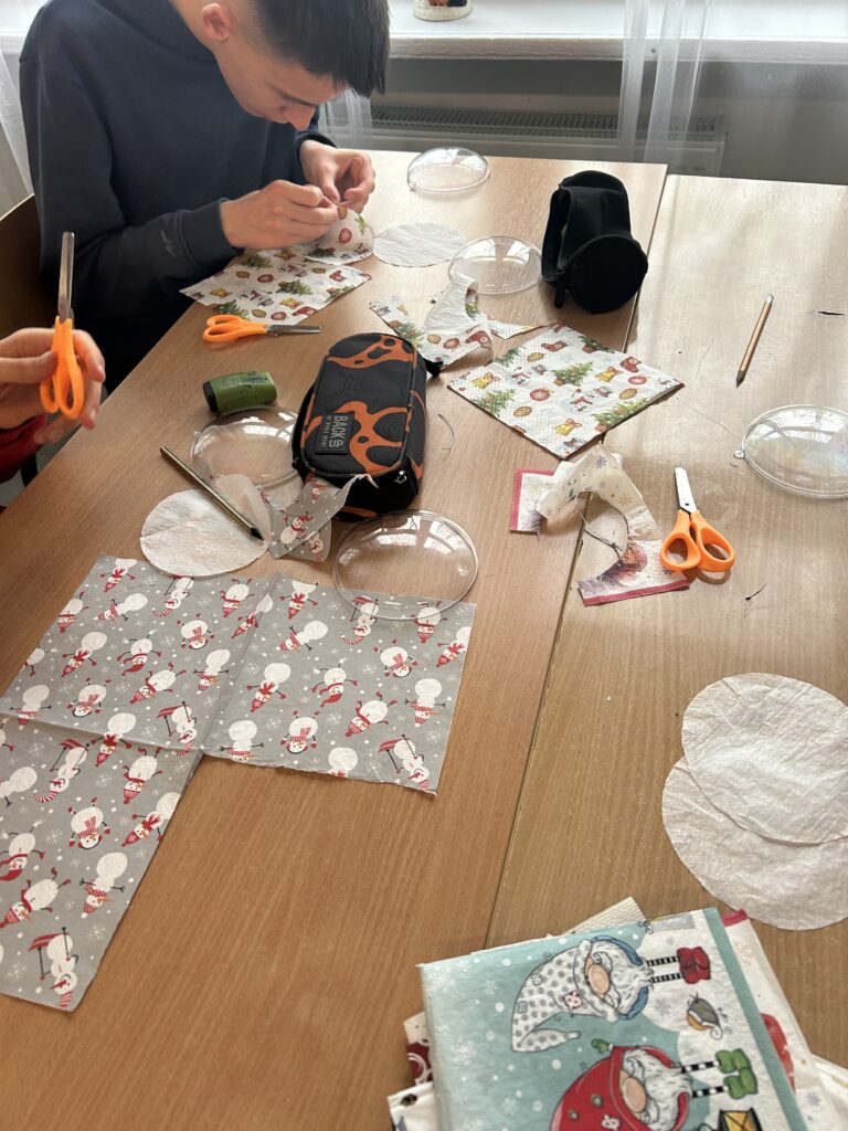 na ławkach leżą serwetki, nożyczki, piórnik. Chłopiec skupiony na rozwarstwianiu serwetek, którymi będą wyklejane plastikowe bombki