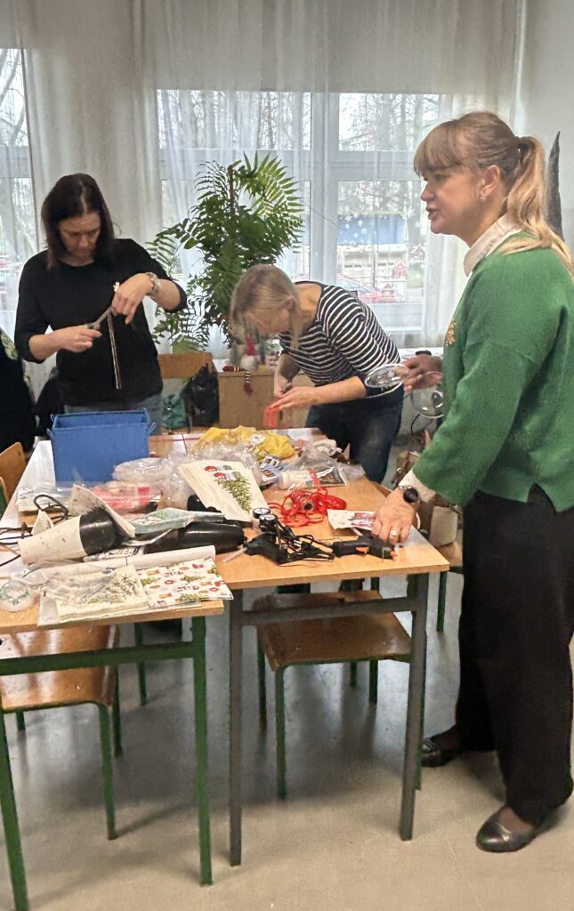 na ławkach różne materiały. Przy ławkach stoją 3 panie. Jedna wyjaśnia dzieciom, co mają robić, dwie pozostałe przygotowują tasiemki do wyklejani