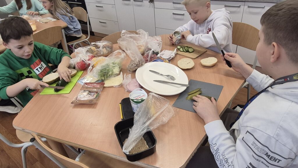W Kuchcikowie stolik zastawiony talerzykami, foliowymi torebkami z jedzeniem. Uczniowie mają przed sobą deski do krojenia i kroją ogórki.