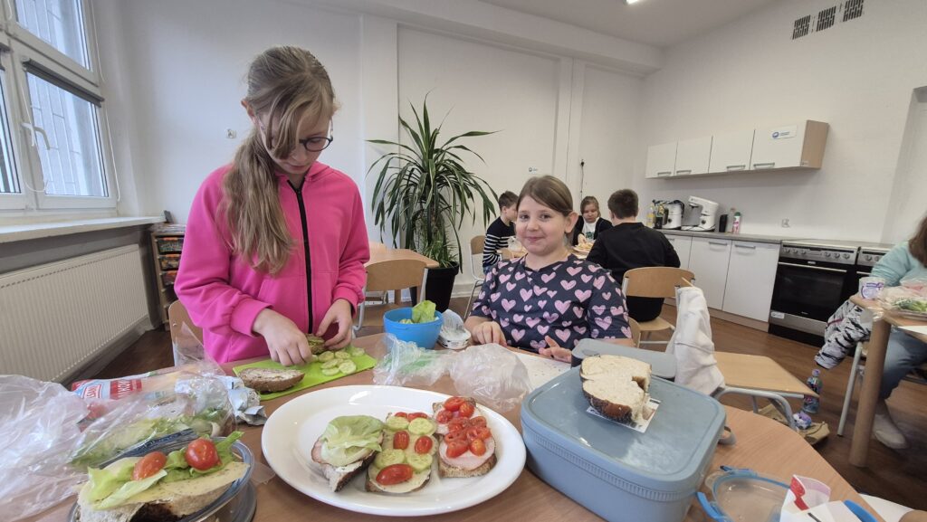 Na pierwszym planie stolik z talerzem z kolorowymi kanapkami, w tle uśmiechnięta uczennica.