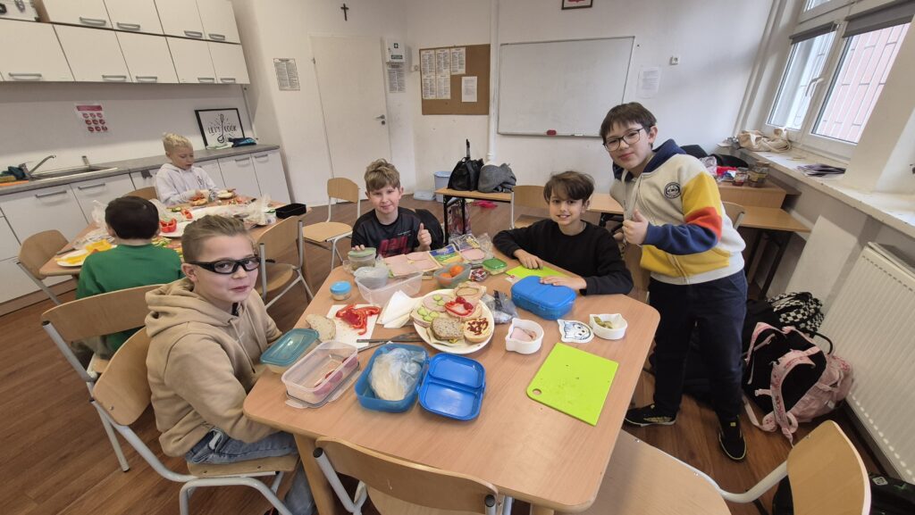 W Kuchcikowie uczniowie IVa siedzą przy stole zastawionym produktami spożywczymi, po środku stołu talerz pełen kolorowych kanapek.