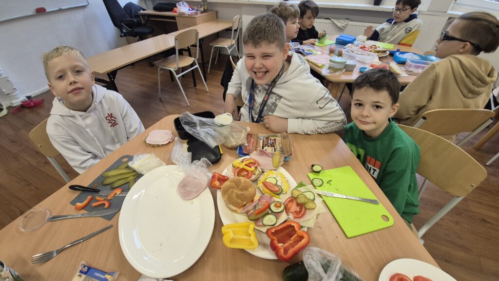 Uczniowie IV a siedzą przy stole zastawionym produktami spożywczymi, po środku stołu talerz pełen kolorowych kanapek.