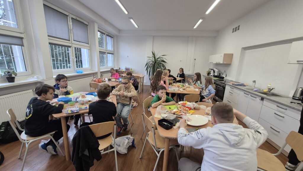 Uczniowie IVa w Kuchcikowie siedzą przy pięciokątnych stolikach, na których znajdują się produkty spożywcze i talerzyki.  