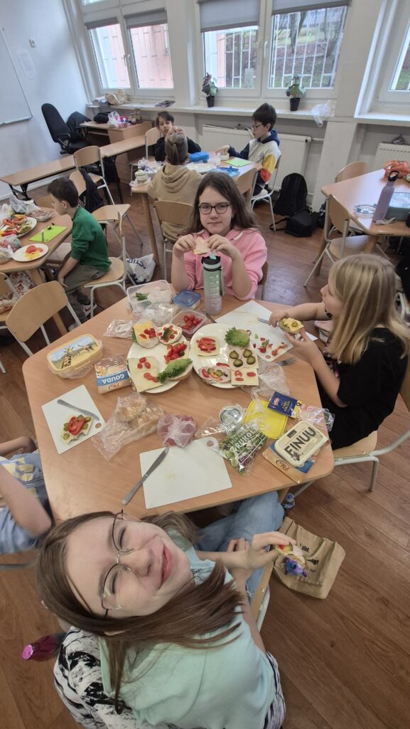 W Kuchcikowie na pierwszym planie uśmiechnięta uczennica IVa, dalej stolik zastawiony produktami spożywczymi i dwa talerze pełne kolorowych kanapek, którymi częstują się dwie uczennice.