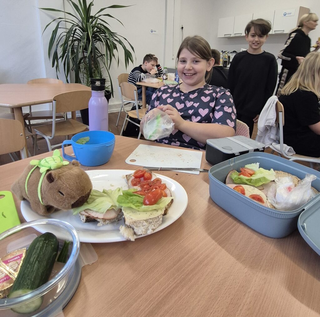 W Kuchcikowie na pierwszym planie stolik z talerzem z kolorowymi kanapkami, w tle uśmiechnięta uczennica.
