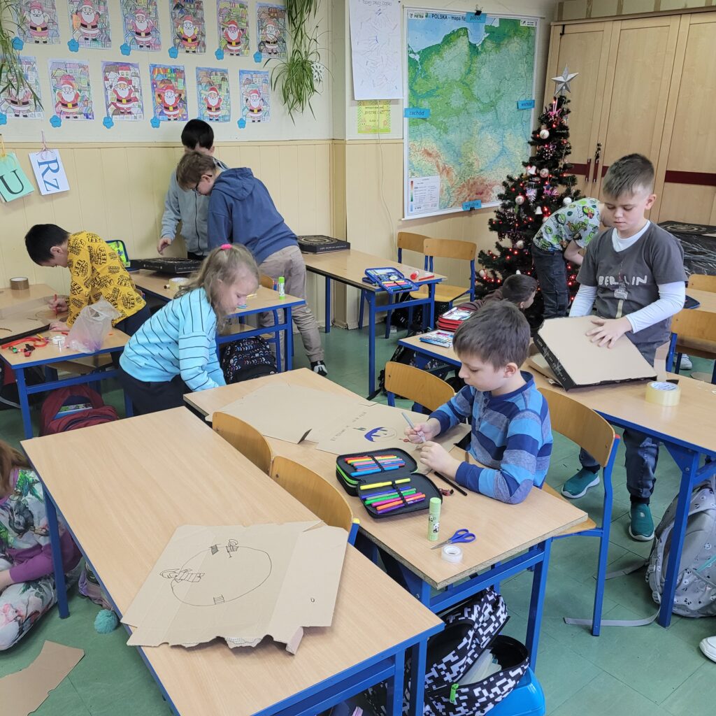 dzieci pracują na podłodze i przy ławkach. Malują na pudełkach po pizzy. 