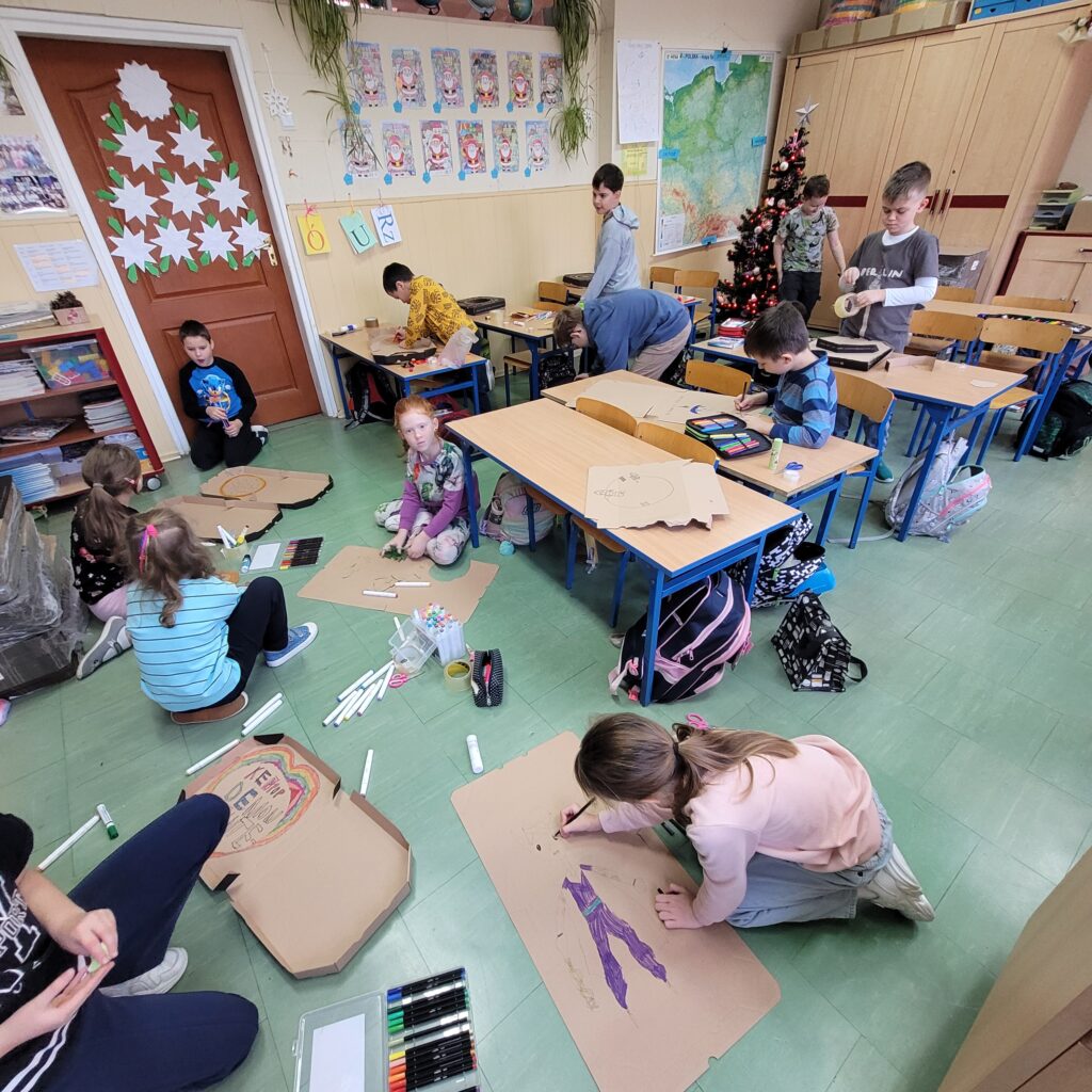zdjęcie widoku klasy. Dzieci siedzą na podłodze lub przy ławkach. Malują i wycinają.