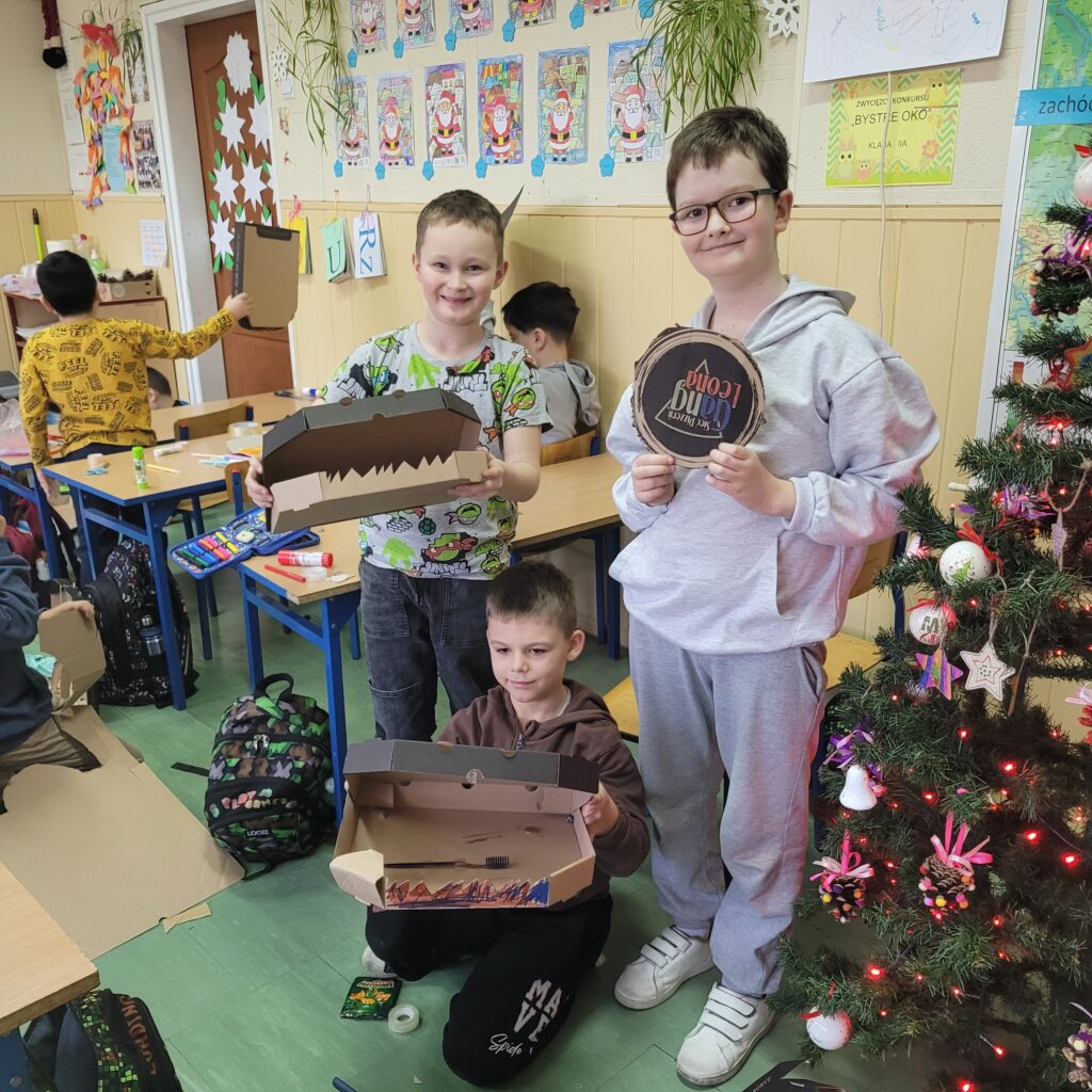 widzimy grupę uczniów trzymających swoje kolorowe prace wykonane z pudełek po pizzie