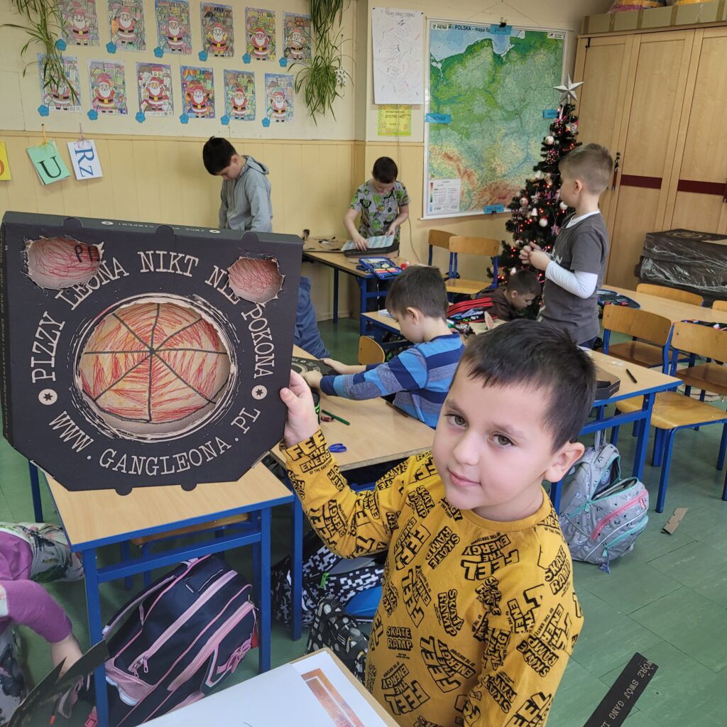 uczeń trzyma pudełko po pizzie ozdobione przez siebie, widać dwie dziury w pudełku